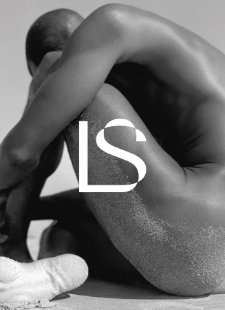 Black and white image of a nude adult male sitting on a sandy beach with large white text overlayed. The background is a bright sky.