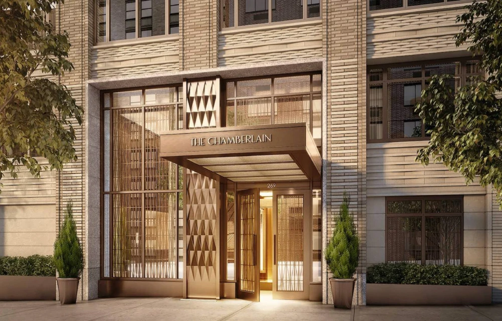 Exterior of The Chamberlain building with a patterned entrance, potted evergreen plants, and a light brick facade in an urban setting.