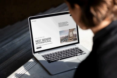 Over-the-shoulder view of a person looking at an open silver laptop displaying a Nest Seekers International website mockup.