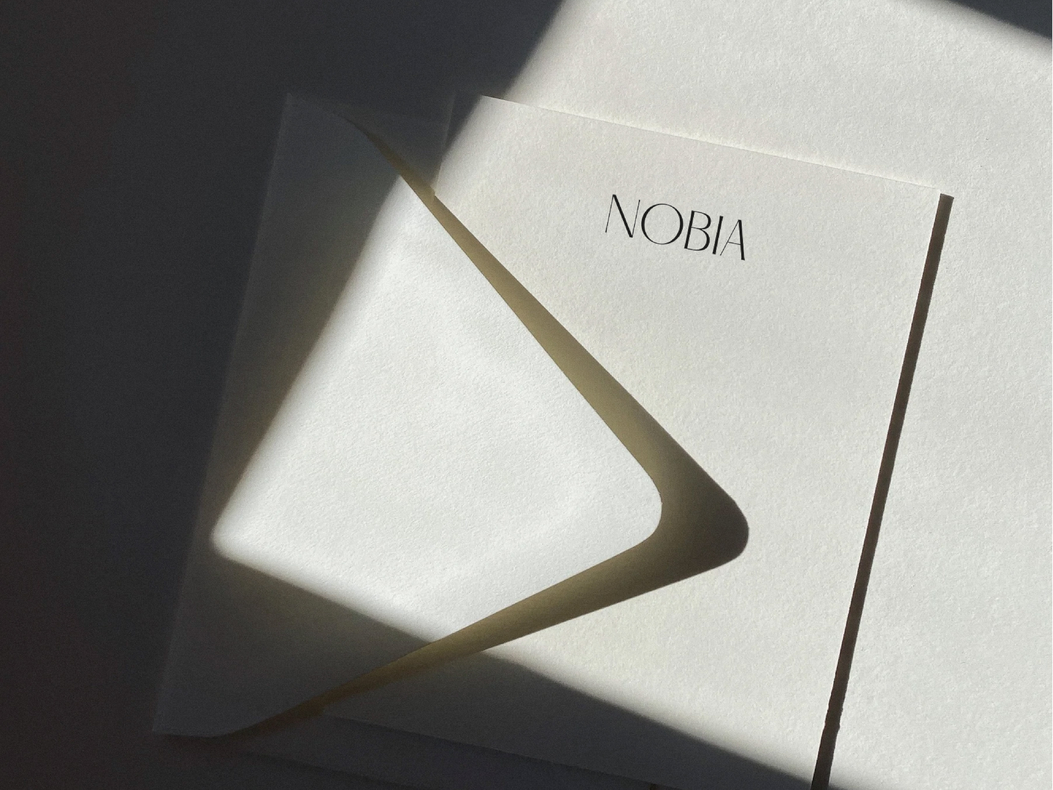 Minimalist composition featuring a white invitation or greeting card with the word NOBIA against a neutral background with light and shadow.