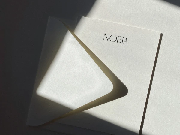Minimalist composition featuring a white invitation or greeting card with the word NOBIA against a neutral background with light and shadow.