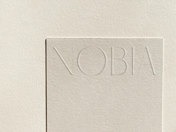 Close-up of a textured, neutral-toned cardstock with the word 'NOBIA' subtly embossed on its surface in simple, sans-serif typography.