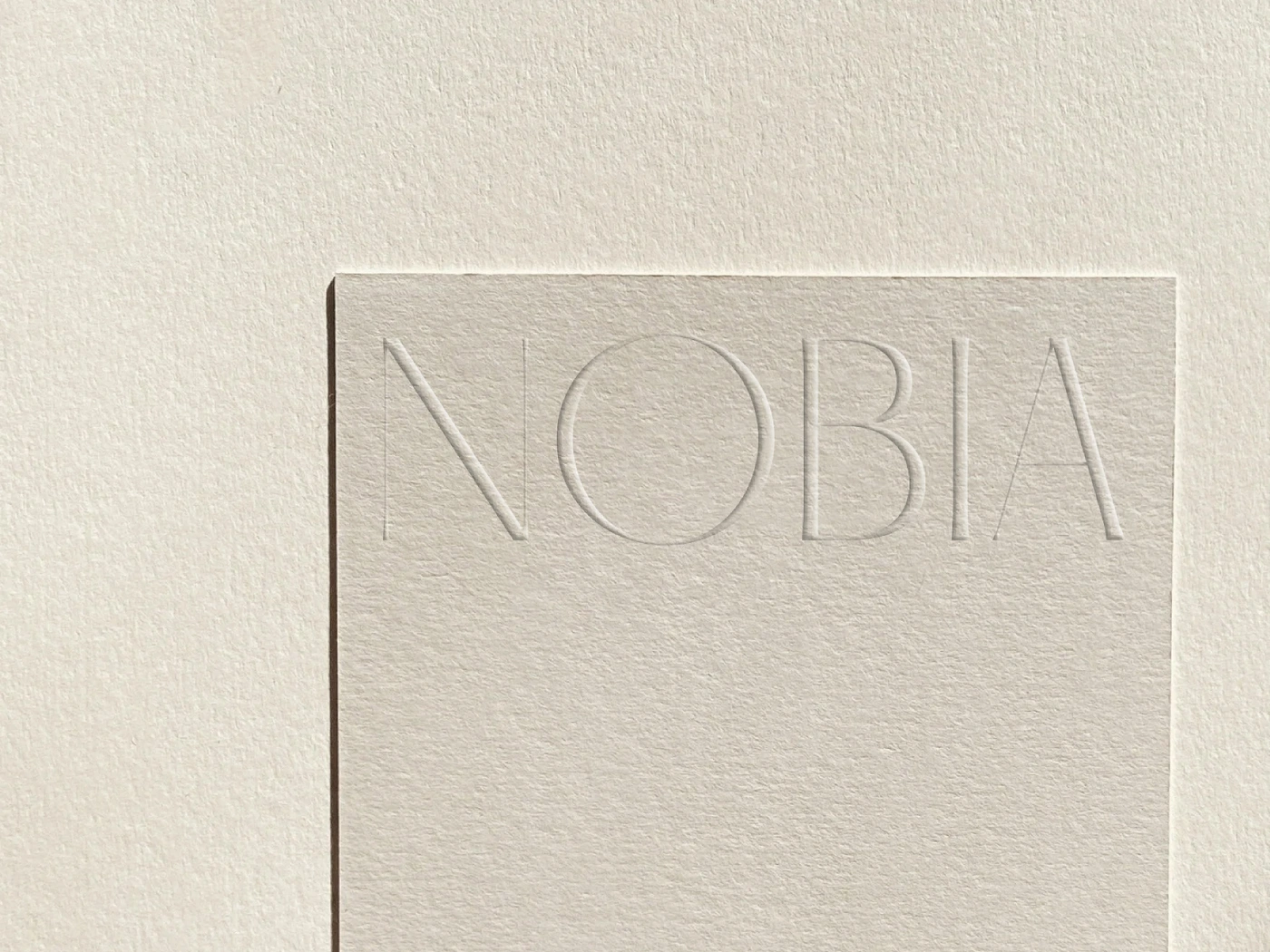 Close-up of off-white textured paper card debossed with the word NOBIA under soft natural directional light.