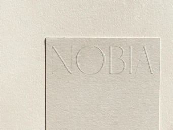 Close-up of off-white textured paper card debossed with the word NOBIA under soft natural directional light.