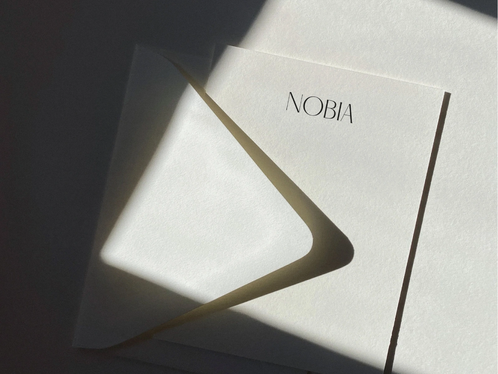 Minimalist off-white stationery card and envelope with black Nobia branding in natural diagonal sunlight and shadow.