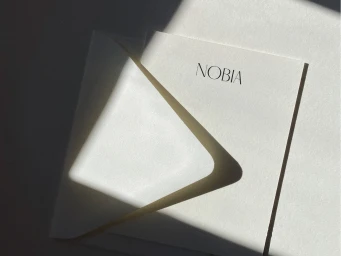 Minimalist off-white stationery card and envelope with black Nobia branding in natural diagonal sunlight and shadow.