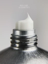 Close-up of white skincare cream dispensing from a brushed silver aluminum tube nozzle against a neutral grey background.