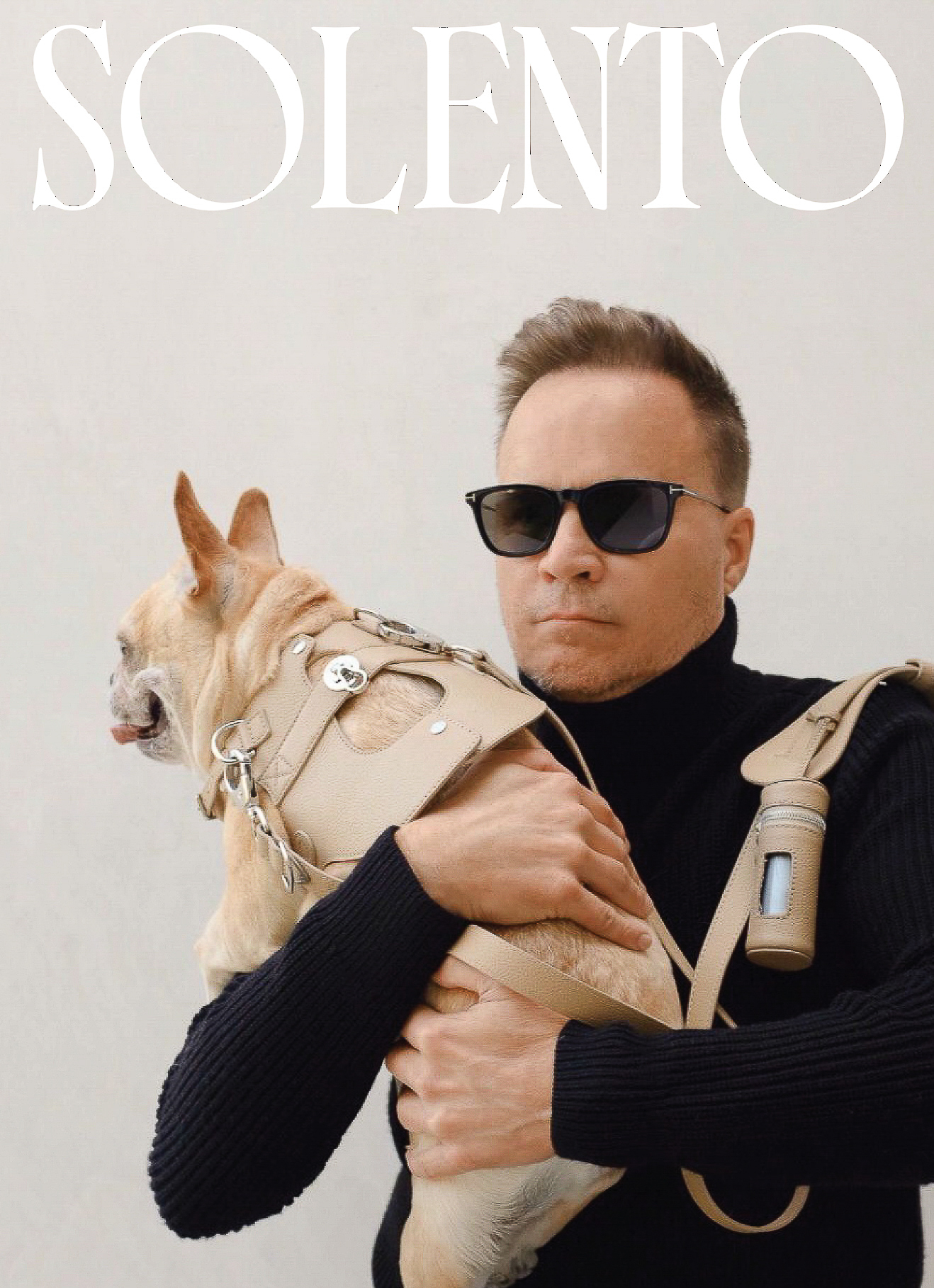 Tan French bulldog wearing tan harness held by an adult man wearing a black turtleneck and sunglasses against a white background.