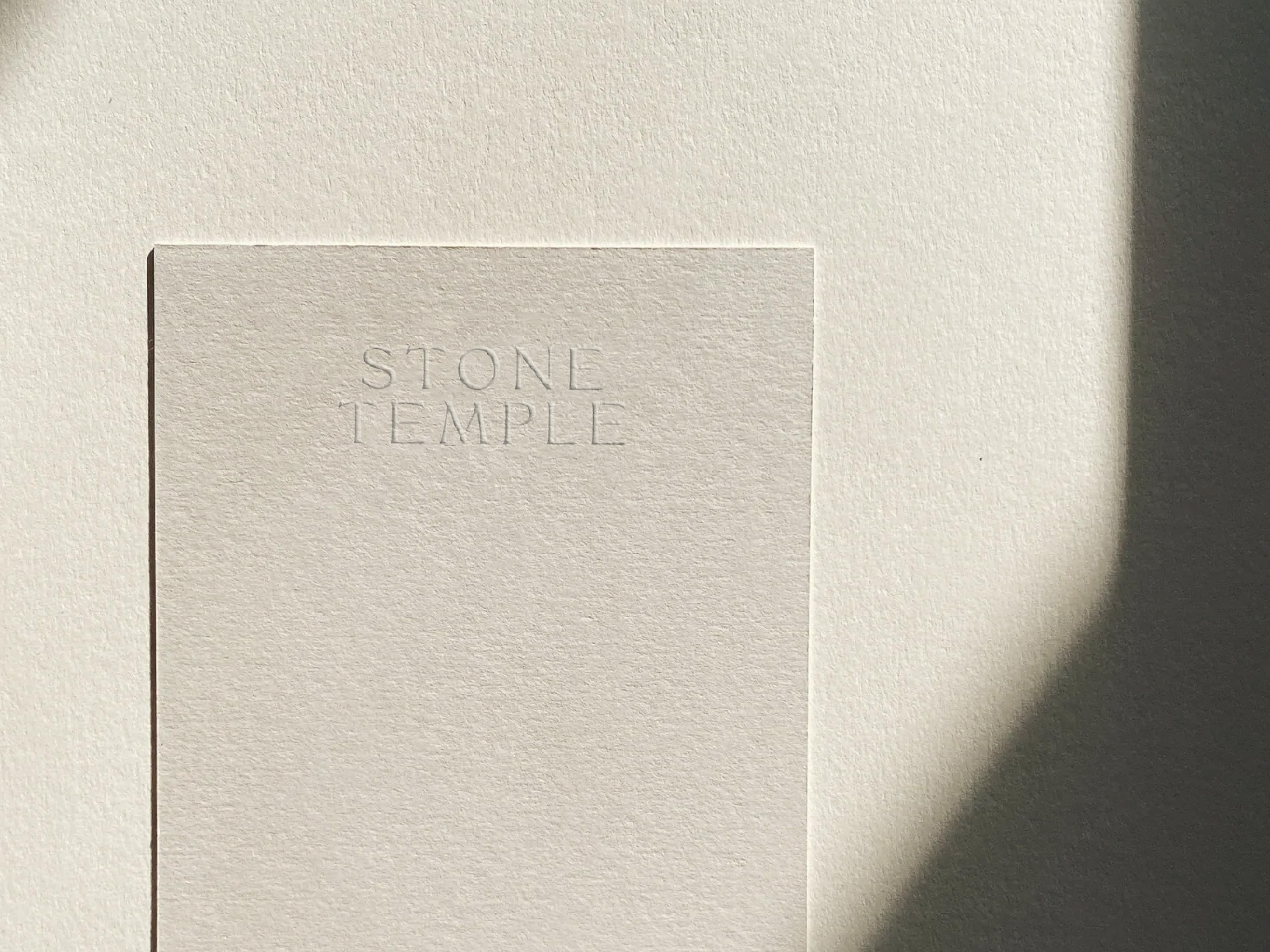 Close-up of a card with the embossed words 'STONE TEMPLE' on a cream-colored textured surface with a gentle light and shadow gradient.