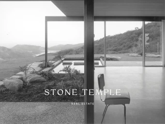 Black and white architectural view through glass walls with a modern chair, reflecting pool, and hillside landscape in the background.