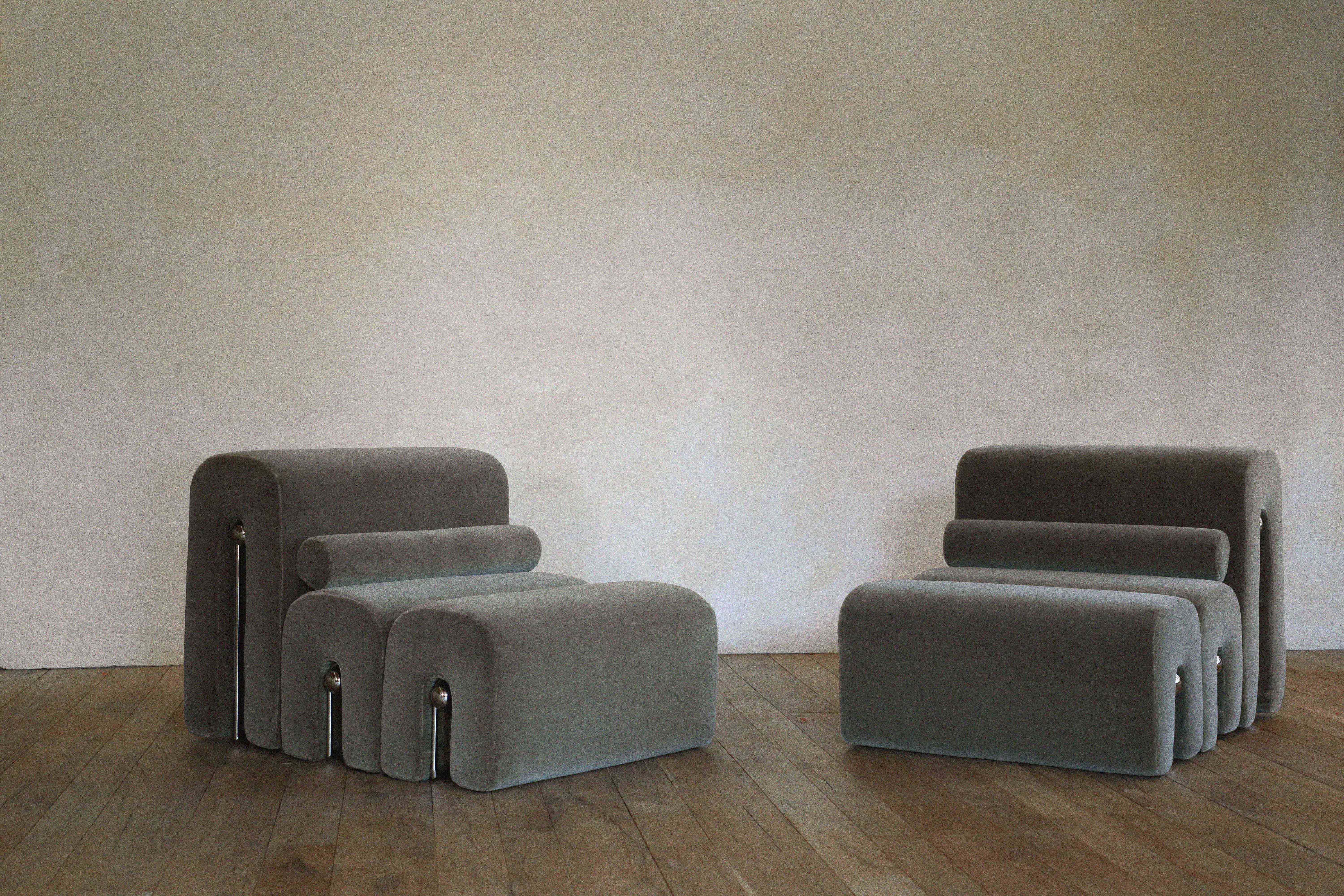 Two modular upholstered grey lounge chairs with tubular cushions resting on a rustic wood floor against a neutral plaster wall.