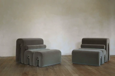 Two modular upholstered grey lounge chairs with tubular cushions resting on a rustic wood floor against a neutral plaster wall.