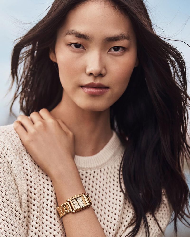 Portrait of a young Asian woman wearing a gold wristwatch and cream-colored sweater, gazing at the camera with natural makeup and long brown hair.
