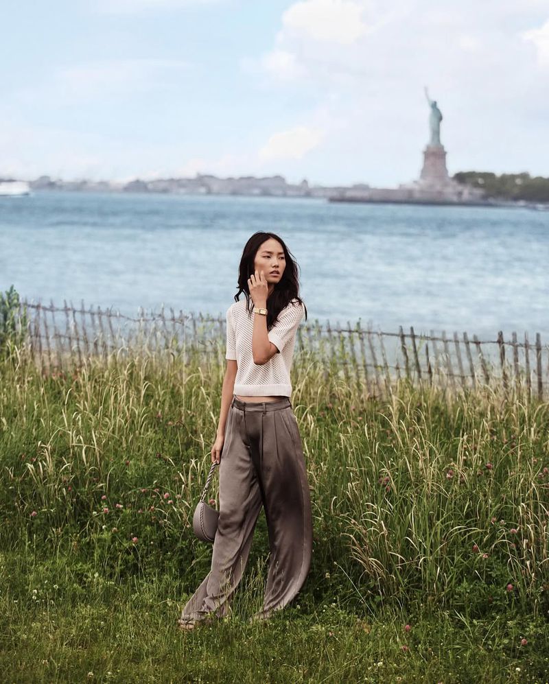 Asian woman stands in a grassy field with the Statue of Liberty in the background, wearing a cream sweater and grey satin pants.