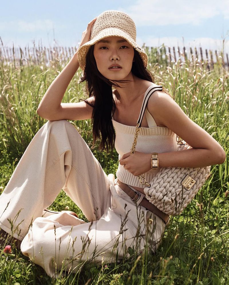 A young Asian woman poses in a grassy field wearing a straw hat, light-colored tank top, and pants, with a chain-strap purse.