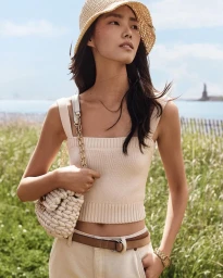 Model Jiahui Zhang in a cream knit tank top and straw bucket hat with a woven chain bag standing in an outdoor grassy field.