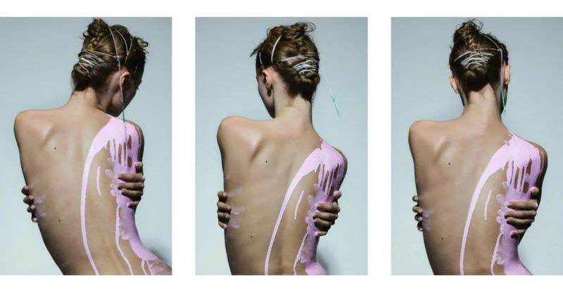 Back view of a slender person with a light skin tone and their arms crossed, with pink paint dripping down their shoulder and back.