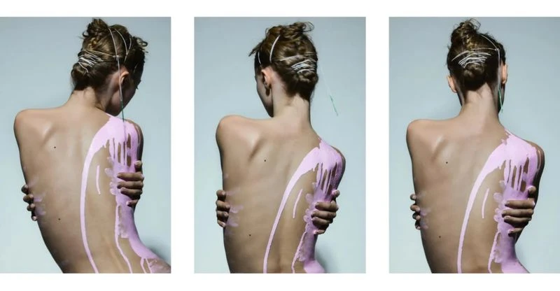 Back view of a slender person with a light skin tone and their arms crossed, with pink paint dripping down their shoulder and back.