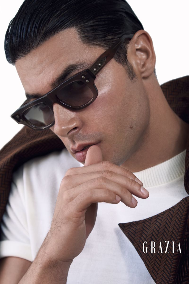Portrait of an adult male wearing brown sunglasses and a white shirt, with a brown jacket draped over his shoulder against a white backdrop.