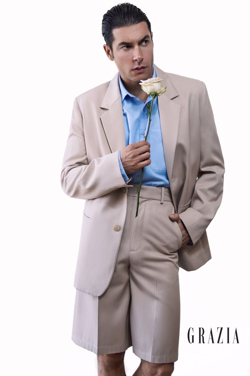 Elegant man in beige suit and shorts holding a white rose, posing in a studio with a clean backdrop for a fashion magazine editorial.