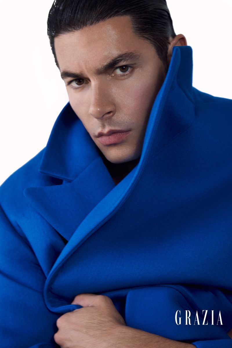 Man in bright blue oversized coat poses in a studio against a white backdrop, looking at the camera in this fashion advertisement.