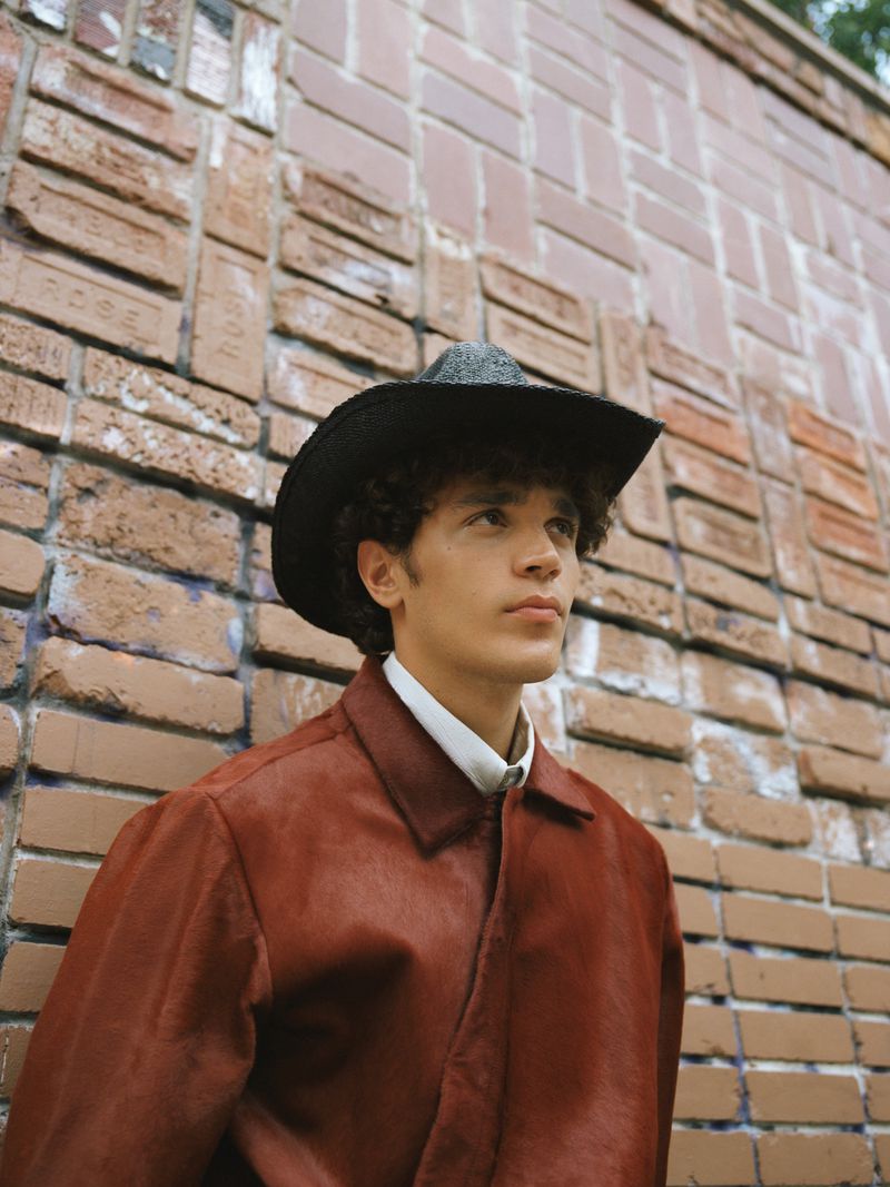 Young adult male in a rust-colored jacket and black cowboy hat leans against a textured brick wall in a medium shot, gazing upward.