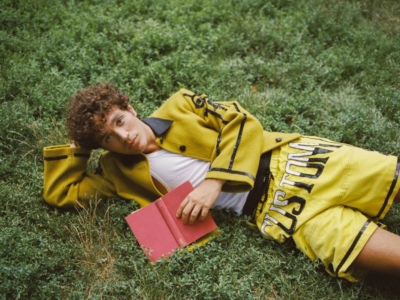 Young adult laying in the grass, wearing yellow jacket and shorts, holding a red book, outdoor setting, medium shot.