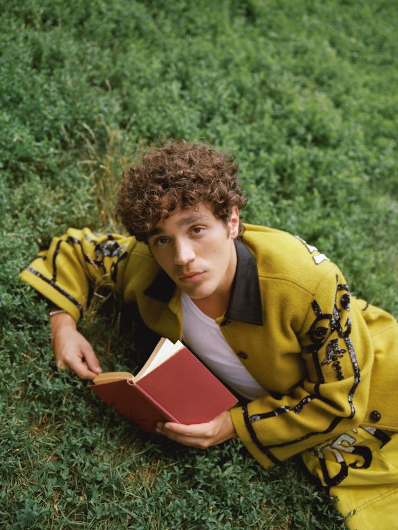 David Iacono lying on a green grassy lawn wearing an embellished yellow jacket and holding an open red book.