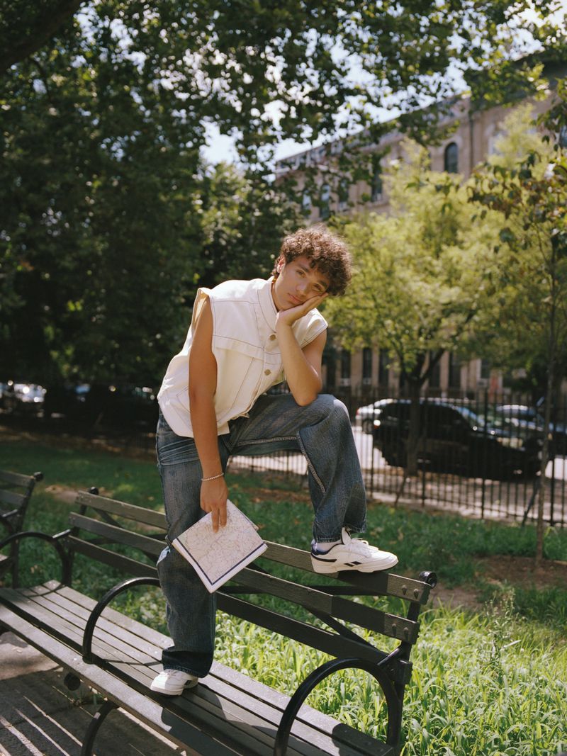 Adult male with curly hair sitting on a park bench wearing a sleeveless top and jeans, holding a notepad in an outdoor setting.