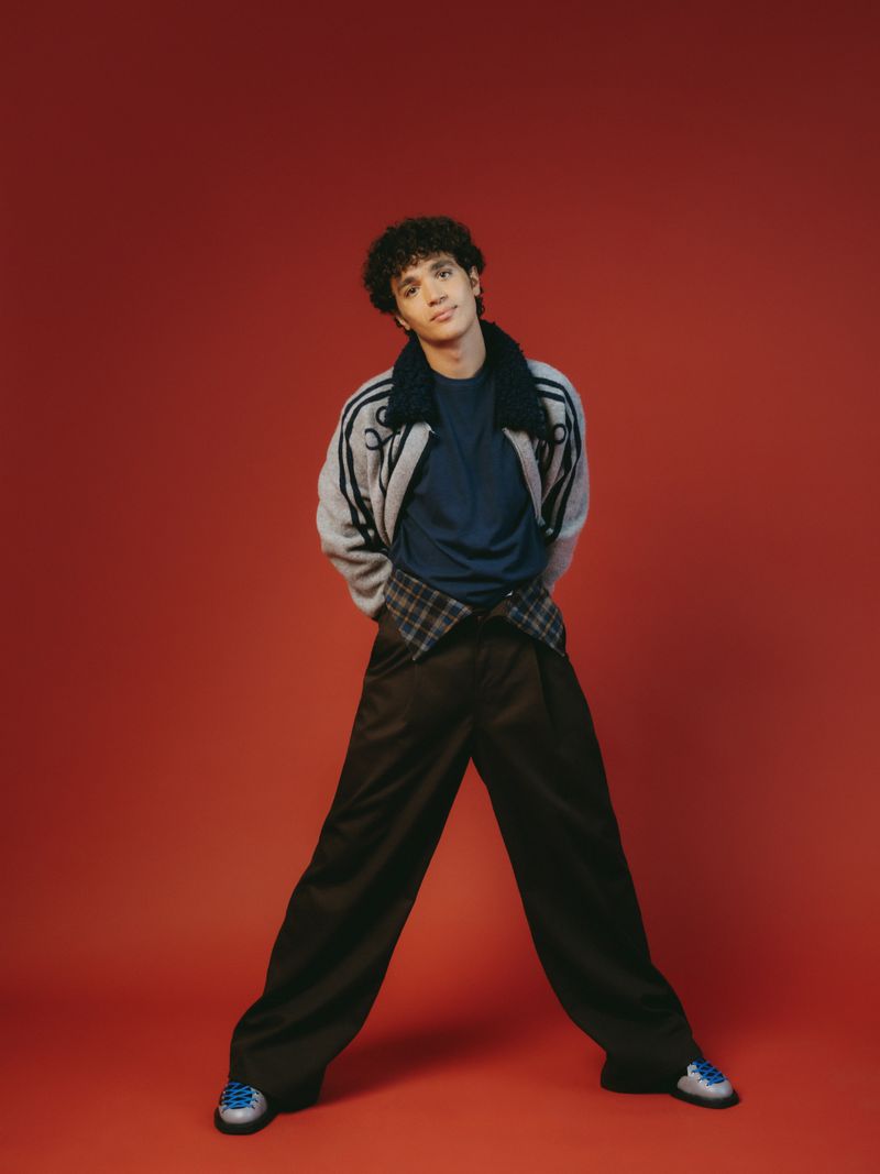 Young adult male stands in a relaxed pose against a red backdrop wearing an oversized jacket, blue shirt, and wide-legged trousers with blue sneakers.