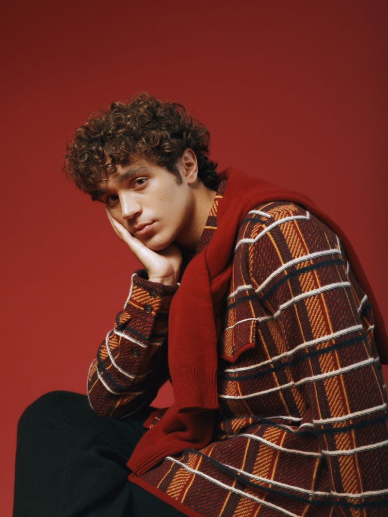 Portrait of a male model with curly hair wearing a plaid sweater and red sweater, against a solid red backdrop in a photo studio.