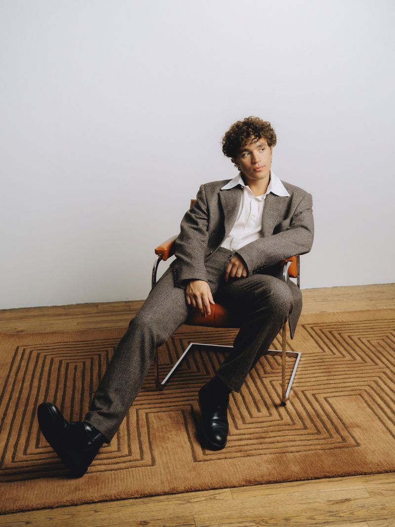Adult male with curly hair in a grey suit and white shirt seated in an orange chair indoors on a brown rug, looking off-camera.