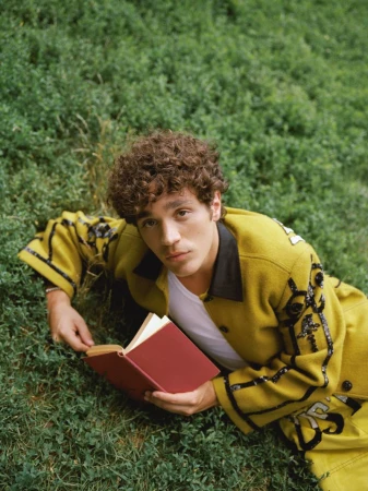 David Iacono lying on a green grassy lawn wearing an embellished yellow jacket and holding an open red book.