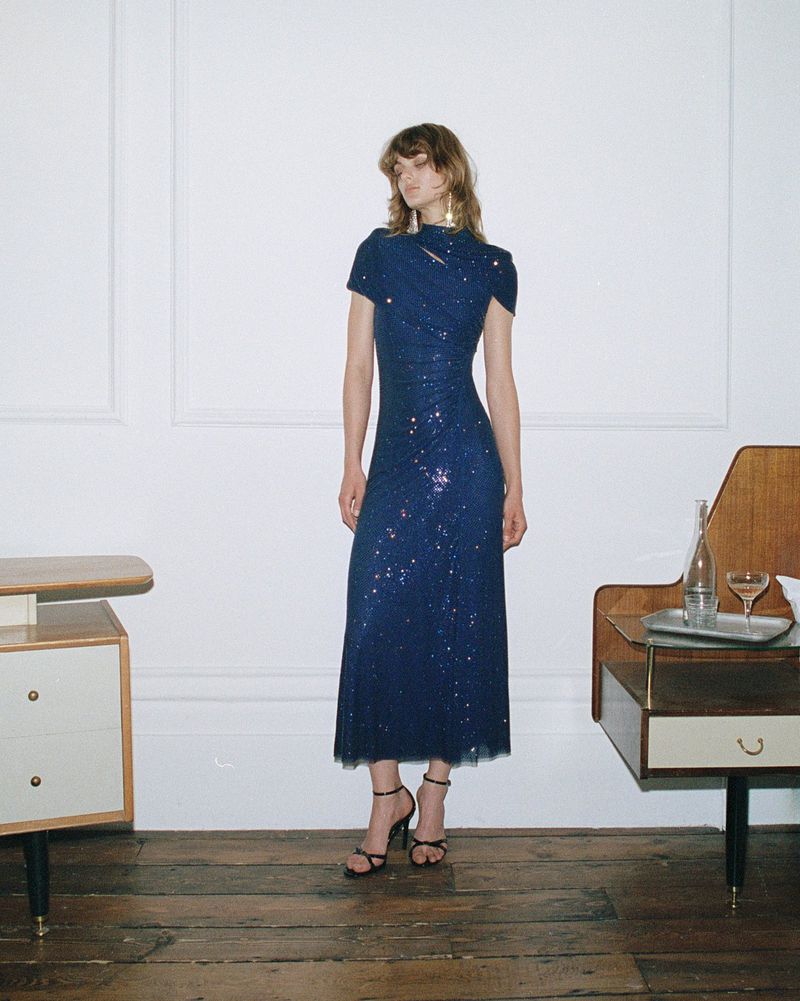 A slender woman with shoulder-length brown hair poses in a form-fitting, sparkling navy midi dress and strappy black heels in a minimalist indoor setting.