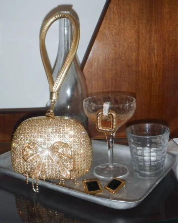 Gold glitter purse with bow and handle, earrings, and coupe glass with drink on a silver tray against a blurred background.