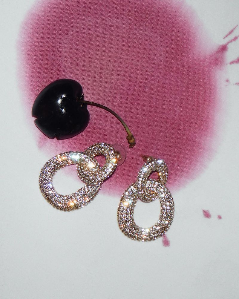 Sparkling earrings next to a cherry on a white surface with abstract red-purple ink blotches. Jewelry and fruit, still life.