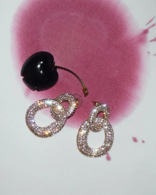 Sparkling earrings next to a cherry on a white surface with abstract red-purple ink blotches. Jewelry and fruit, still life.