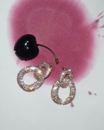 Sparkling earrings next to a cherry on a white surface with abstract red-purple ink blotches. Jewelry and fruit, still life.