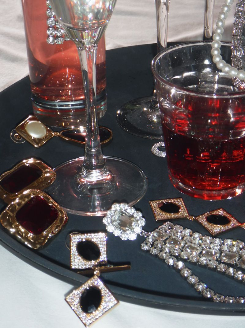 Close-up of glassware filled with red liquid on a black tray, surrounded by jewelry and decorative objects against a neutral backdrop.