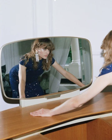 Model Matilda Navratil reflected in a vanity mirror wearing a sparkling navy blue Self-Portrait dress and crystal drop earrings.