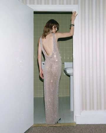 Model Matilda Navratil in a sparkling backless maxi dress posing in a bathroom doorway with green tiles.