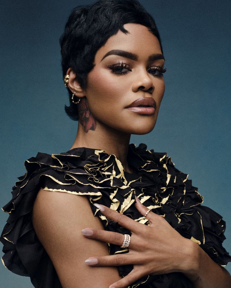 Headshot of an African-American woman with dark hair, wearing a black ruffled top with gold trim against a muted blue background.