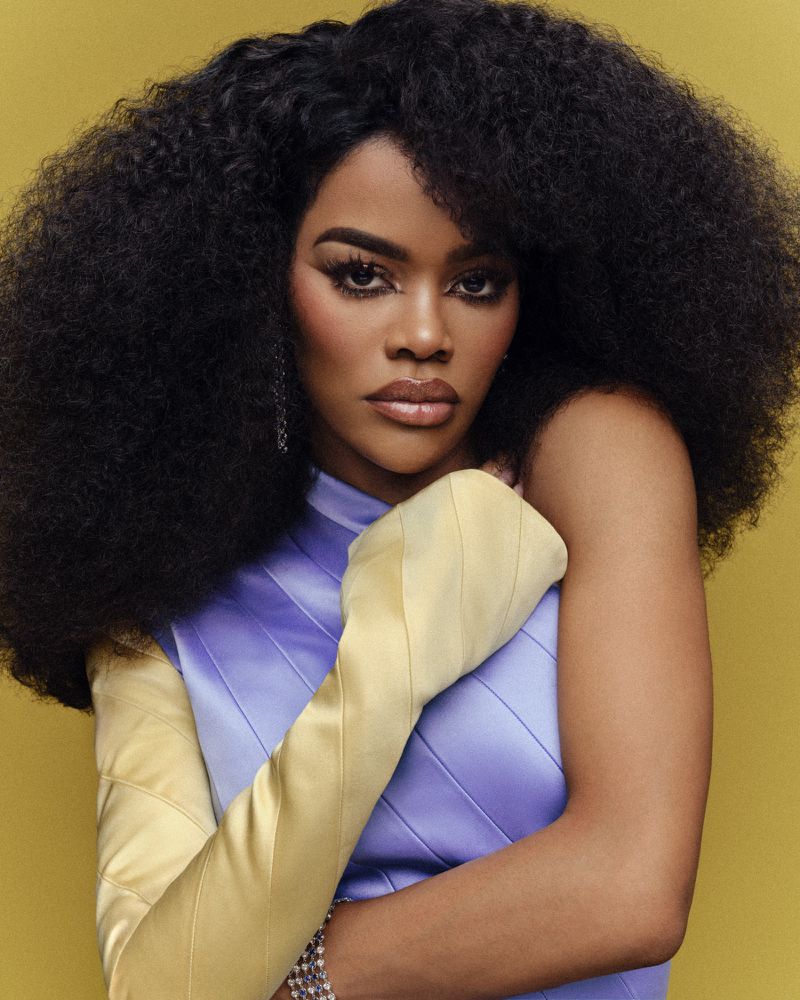 A striking studio portrait of a Black woman with large, afro-textured hair, wearing a lilac dress and long yellow gloves against a yellow backdrop.