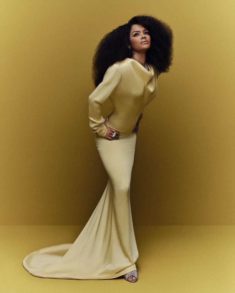 Elegant African-American woman in a draped golden gown strikes a pose in a studio setting, showcasing fashion and beauty with style.