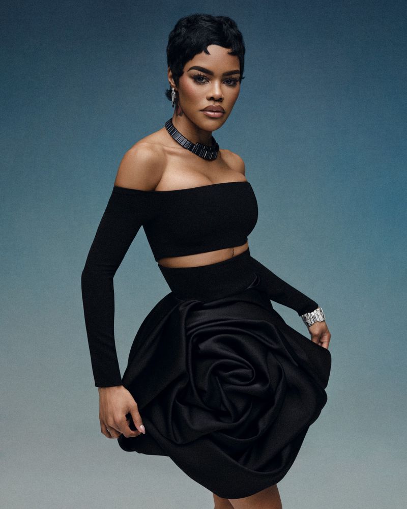 Fashion portrait of a Black woman wearing a black crop top, rose skirt, jewelry, and pixie haircut, posing against a blue-gradient background.