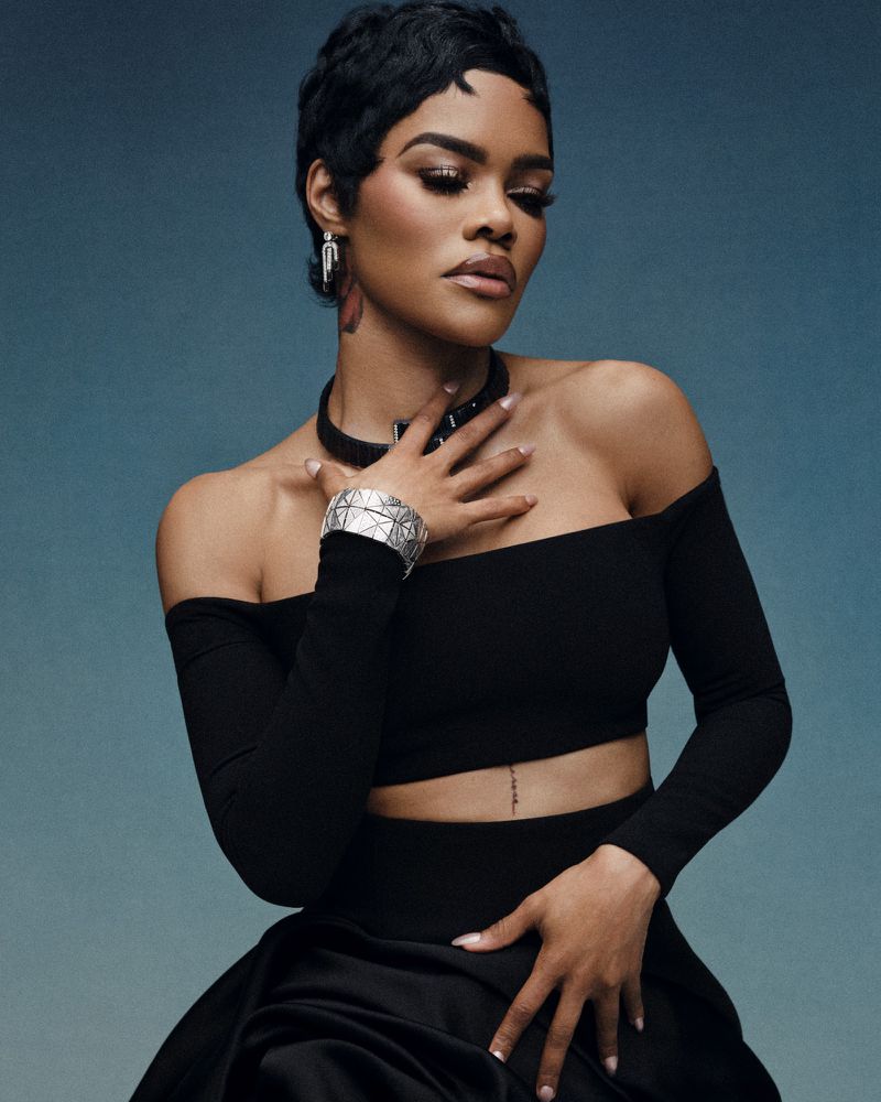Portrait of an adult woman with short dark hair in an off-the-shoulder black top, a choker, and a large bracelet, posing indoors.