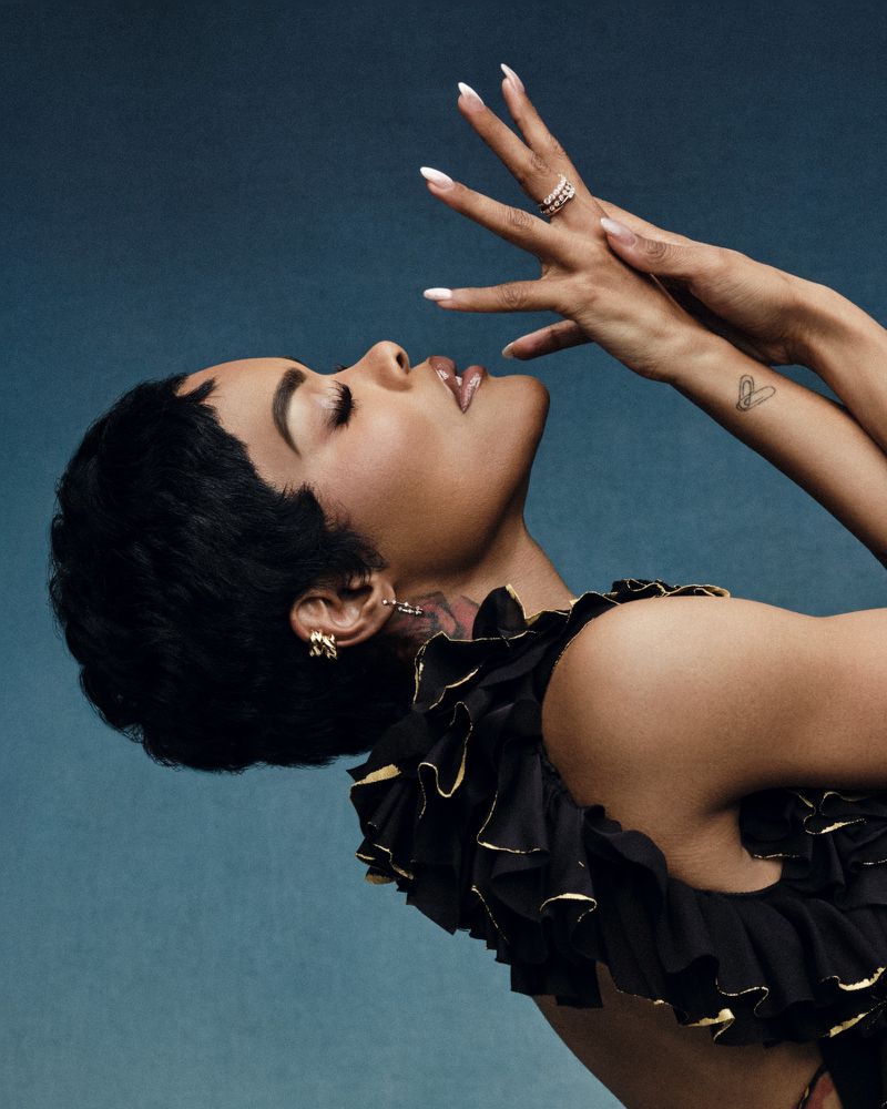 Portrait of an adult woman with black ruffled top and short hair looking up with hands near her face in a blue setting.