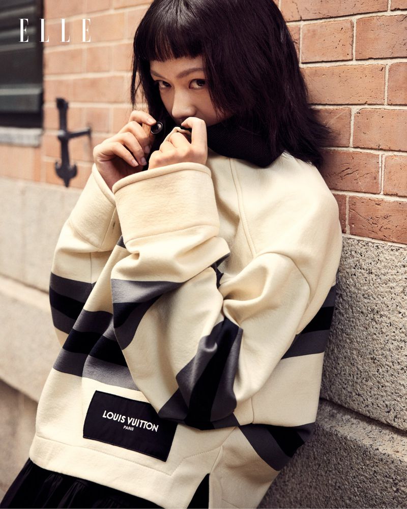 Elegant Asian woman in a Louis Vuitton ivory sweater poses by a brick wall for an ELLE magazine fashion editorial.