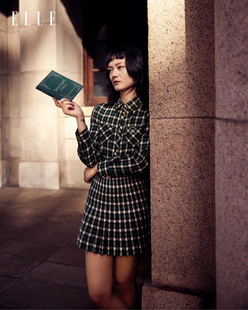 Young woman in a plaid dress holds a book standing near a stone column and window. Magazine logo visible. Fashion editorial style photo.