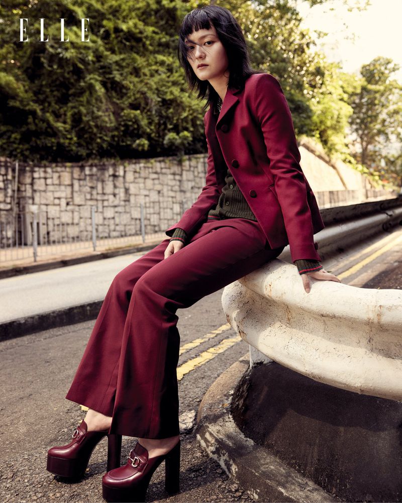 Editorial fashion shot of Asian woman in a burgundy suit and platform shoes, sitting on a roadside barrier with greenery in the background.
